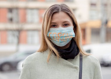 Corona virüsünden korumak için maskeli bir kız. Maskede Corona yazıyor. Klinik binasının önünde duran maskeli kadın. Tıbbi maskeli güzel sarı saçlı kız. Corona virüsü salgını