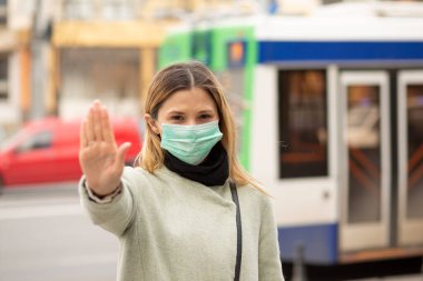 Steril koruyucu tıbbi maske takan genç bir kadın doktor, onu durduruyor. El işaretleriyle işaret veriyor. Şehrin arka planında virüse hava yoluyla yakalanmayı reddeden biri yok.