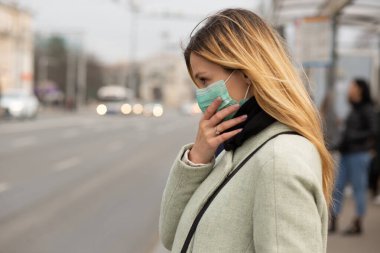 Beyaz kadın steril koruyucu tıbbi maske takıyor. Covid-2019 Corona virüsü taşıyormuş. Avrupa 'da bir caddenin ortasındaki otobüs durağında. 