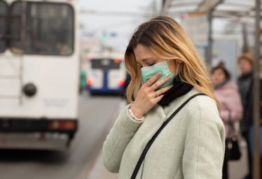 Sad Caucasian woman wearing sterile protective medical mask ill by the Covid-2019 Corona virus at public bus station in a European city street feeling bad looking down to the side people on background clipart