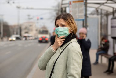 Avrupa 'nın bir caddesindeki otobüs durağındaki Covid-2019 Corona virüsünün etkisinde steril koruyucu tıbbi maske takan üzgün beyaz kadın kameralara bakarken kendini kötü hissediyor.