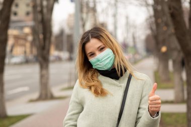 Güzel bir kız, yüzünde steril koruyucu maske takmış genç bir kadın, Avrupa 'da bir şehirde yürüyor, baş parmağını kaldırıyor, jest yapıyor gibi. Çevresel ekoloji kirliliği sorunu Gezegen konseptimizi kurtarır
