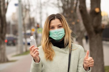 Hasta, mutlu, neşeli bir kız, Coronavirus tarafından korunan ameliyat maskesi takan sarışın genç bir kadın, Covid2019 el dezenfektanı gibi el ele tutuşarak baş parmak hareketi yapıyor.