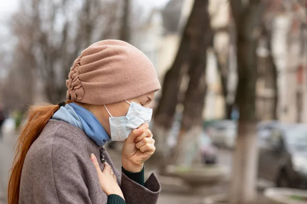 Sick girl, a redhead young in profile woman coughing wearing surgery medical mask feeling bad, ill, being hit by the coronavirus, Covid2019 standing outdoors in the city.