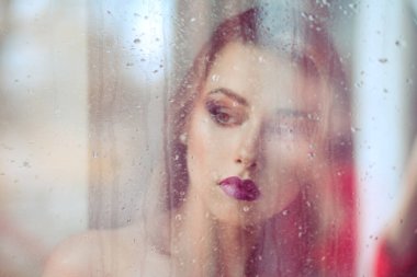 Güzel, melankolik, yalnız, üzgün bir kadın pencerenin önünde duruyor, üzgün, düşünceli bir şekilde aşağı bakıyor. Görüntü pencereden geliyor, pencereye yağmur yağıyor ve dışarıdaki manzaraya yansıyor. 