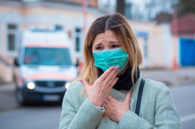 Hasta kız, ameliyat maskesi takan sarışın genç bir kadın kötü hissediyor, hasta, koronavirüs tarafından vuruluyor, Covid2019 Avrupa 'da dışarıda duruyor, arka planda ambulans kliniği var.