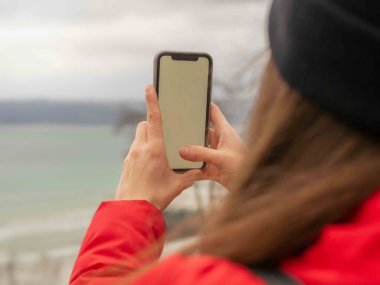 Kırmızı ceketli ve siyah şapkalı bir kadın elinde boş ekranlı bir cep telefonu ile kışın önünde duran doğa grubunun fotoğrafını çekiyor..