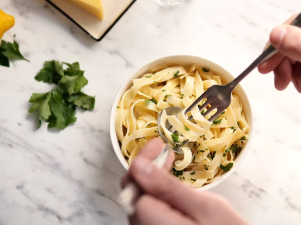 Male hands eating pasta with parsley with fork and spoon from a white bowl on mottle stone table. Top view from above, flat lay. Horizontal image with selective focus