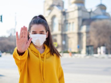 Steril koruyucu tıbbi maske takan genç bir kadın doktor, onu durduruyor. El işaretleriyle işaret veriyor. Şehrin arka planında virüse hava yoluyla yakalanmayı reddeden biri yok.