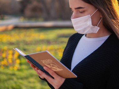 Yan profilli maskeli güzel bir kadın evinin arka bahçesinde geziniyor. Mesafe koyma ve karantina kavramı. Karantina sırasında maskeli genç bir kadın evde kitap okuyor.