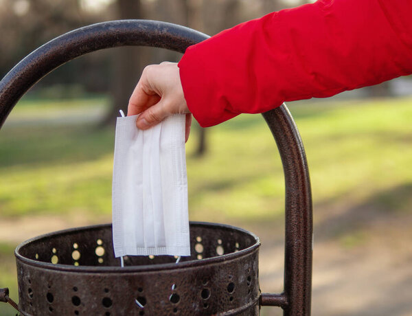 Someone's hand in red coat is throwing the disposable surgical mask into the black rubbish bin in the park. you can not reuse the mask, you have to throw the used mask away in 2 hours concept.