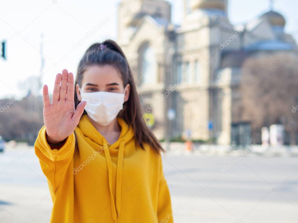 Young female doctor woman wearing sterile protective medical mask, making stop it there sign gesture with hand say signaling No standing on urban city background refusing to catch virus disease by air