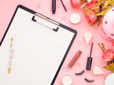 Pano ve makyaj aletleri ve kozmetik ürünleri ve etrafındaki güller pembe renkli arka planda izole edilmiş. Düz konum, üst görünüm kopyalama alanı. Kadınsı güzellik blogcusu iş yeri kavramı.
