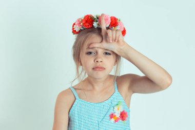 Closeup portrait young unhappy kid girl giving loser sign on forehead looking at you disgust on face isolated on white light green wall background. Negative human emotion face expression body language clipart