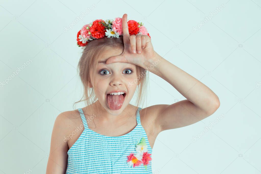 Closeup portrait young unhappy girl, kid giving loser sign on forehead looking at tongue out isolated on light green wall background. Negative human emotion facial expression body language reaction