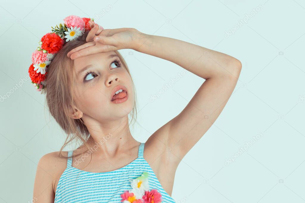 A cute young blond long-haired girl kid standing isolated against a blue-white background while putting two fingers on the forehead with the tongue out looking up, floral headband on head