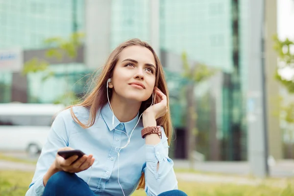 Listening to music audio book. Closeup business woman blue shirt sitting on grass hand on earphones having a conversation on phone looking up thinking how to solve the problem outdoors outside in city