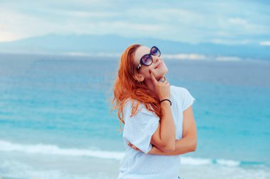 Rahatlatıcı sahil kadını gülümsüyor, mutlu bir şekilde yan tarafa bakıyor. Güzel kız mavi suyla kumsalda hayat hakkında hayal kuruyor. Asya kökenli Çinli ve Kafkas kadın modeli..