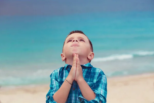 A little boy praying with hands clasped against the backdrop of a stormy sea.