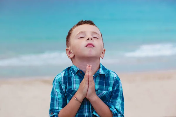 A little boy praying with hands clasped and eyes closed against the backdrop of a stormy sea