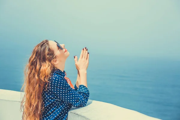 Young woman praying with hands clasped looking up to the sky isolated on seascape ocean background. Travel concept vacation holiday