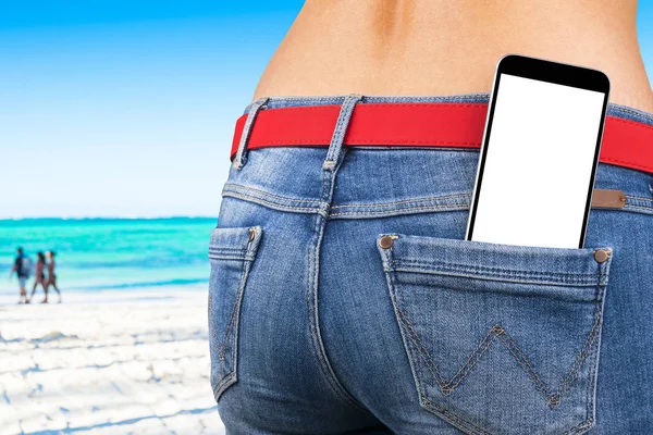 Smartphone con schermo bianco isolato con spazio vuoto per il testo in una tasca jeans di una giovane ragazza snella, sfondo oceano — Foto Stock