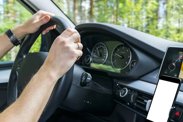 Male hands holding car steering wheel. Hands on steering wheel of a car driving near the lake. Man driving a car inside cabin. Smartphone in holder with  isolated white empty blank screen. Copy space