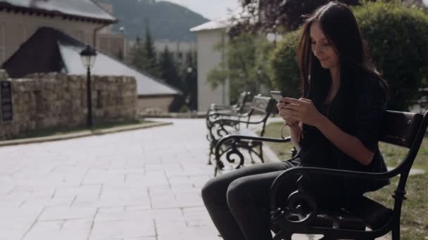 Jovem usando telefone inteligente enquanto ela se senta em um banco no parque da cidade. Ela acena para sua amiga e sai — Vídeo de Stock