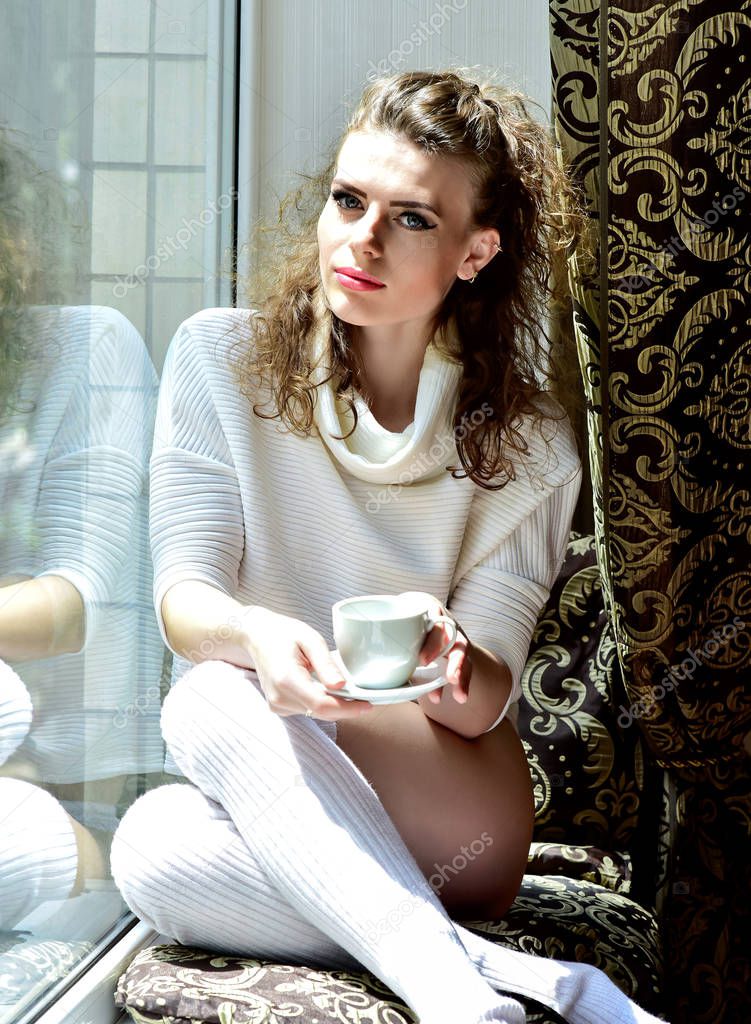 beautiful woman sits by the window on the windowsill in a white sweater in warm white stockings holds a cup in her hands