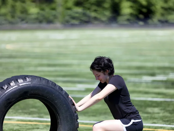 Teen girl pushing heavy old tire on sports field during hot day — Stock Photo, Image