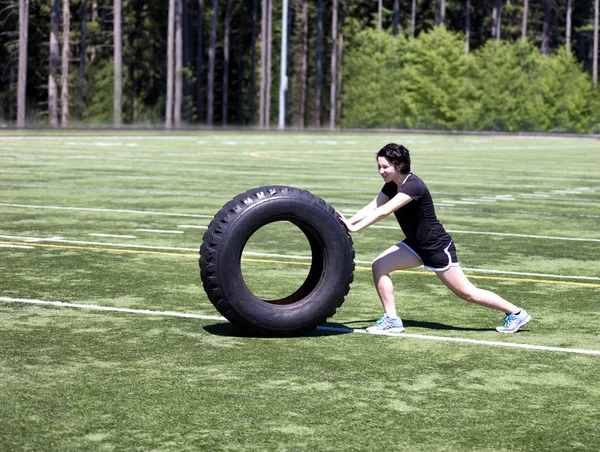 Adolescente chica empujando pesado neumático en el campo de deportes durante el día caliente — Foto de Stock