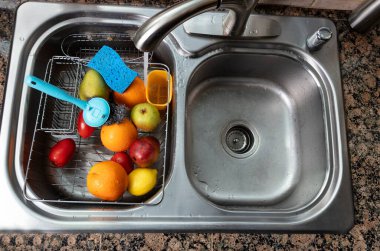 Taze sebzeler ve meyveler mutfak lavabosunda yıkılıyor. Birincil odak noktası musluktan akan su olan yiyecekler..  