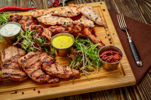 Assorted delicious grilled meat and vegetables with fresh salad and bbq sauce on cutting board on wooden background. Big set of Hot Meat Dishes