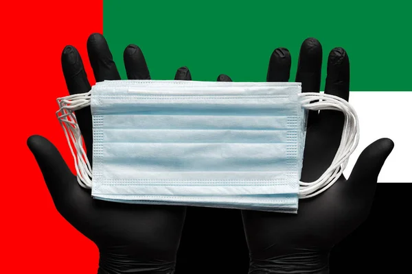 Doctor holds medical face mask in hands in black gloves on background national flag United Arab Emirates UAE. Concept pandemic insurance coronavirus