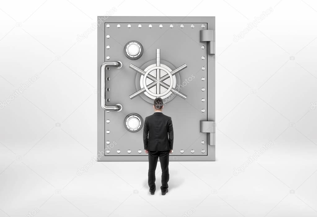 Businessman standing back to us looking at the giant safe door, all isolated on the grey background.