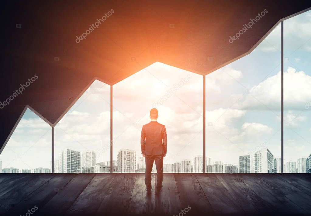 Businessman standing on the floor back to us looking through the big window with cityscape and sunset behind it.