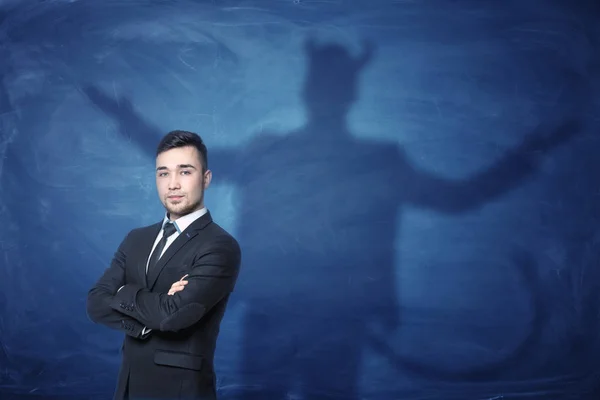 Uomo d'affari in piedi con le mani attraverso e la sua ombra sulla lavagna blu dietro di lui coda come diavolo — Foto Stock