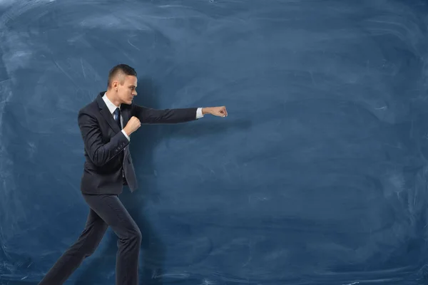 Empresário de terno preto em pé como se ele fosse socar algo invisível perto do quadro azul . — Fotografia de Stock