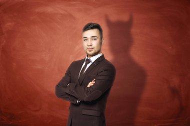 Businessman in suit standing with his arms folded, he is casting shadow of the devil on the rusty orange wall behind him. clipart