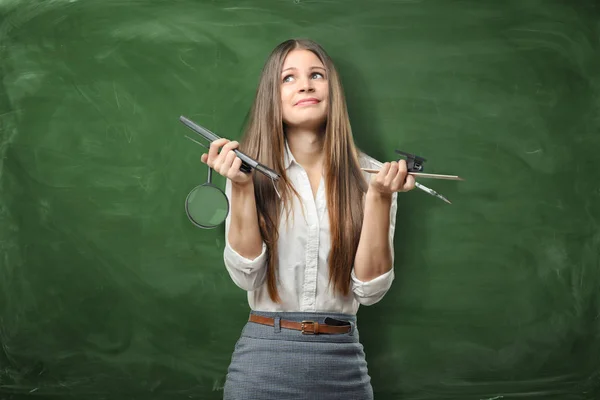 Verward zakenvrouw op groene schoolbord achtergrond bedrijf pennen, liniaal en vergrootglas in haar handen. — Stockfoto