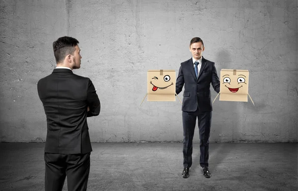 Businessman with a turned back on concrete background looking at another man holding two boxes with drawn faces. — Stock Photo, Image