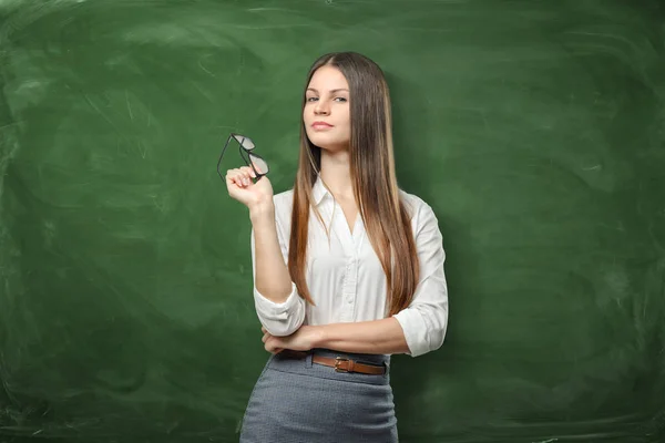 Jonge mooie vrouw houdt haar bril op groene schoolbord achtergrond — Stockfoto