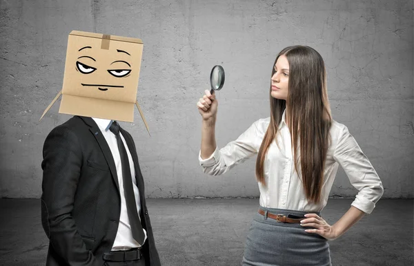 La donna sta guardando attraverso una lente d'ingrandimento un uomo d'affari con una scatola in testa con una faccia da poker — Foto Stock