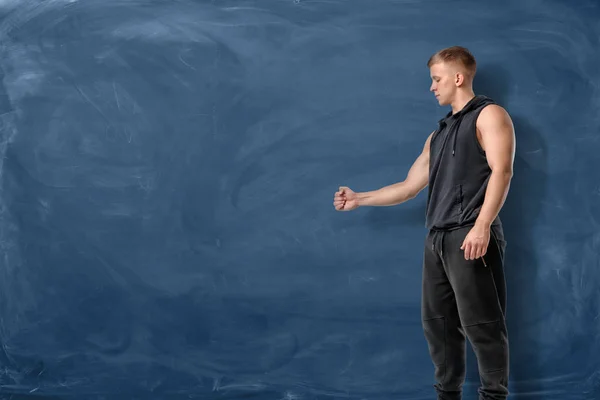 Muscular young man is stretching his fist before him and looking at it on the blue chalkboard background — Stock Photo, Image