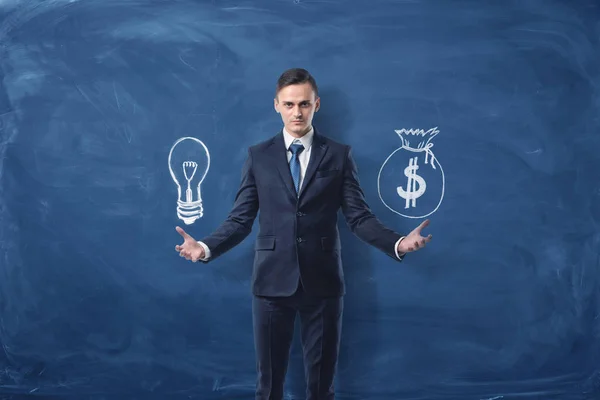 ビジネスマンは、青い黒板背景の電球 1 つの手、別の手でお金の袋を保持しています。 — ストック写真