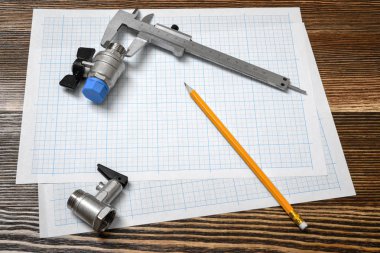 A large vernier scale lying on cross section paper with two ball valves and a pencil lying beside on wooden table background. clipart