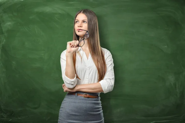 Mooie jongedame is holding haar bril en denken op groene schoolbord achtergrond — Stockfoto