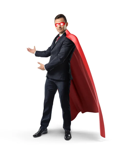 En affärsman i en formell kostym, en röd flödande cape och röda ögon mask visar något bakom honom. — Stockfoto