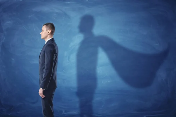 Бизнесмен стоит в профиле и отбрасывает тень плаща суперменов на фоне голубой доски — стоковое фото