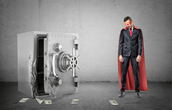 Een triest zakenman in een rode cape en een masker permanent naast een gebroken brandkast met dollarbiljetten rondslingeren. — Stockfoto